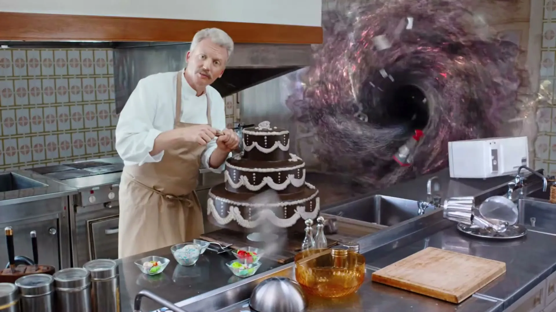 A male chef in a white uniform and apron decorates a chocolate cake in a kitchen, while a swirling vortex appears in the background.