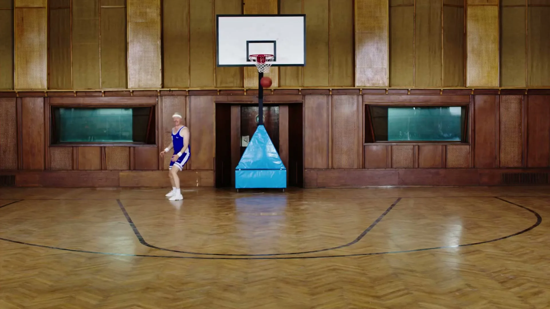 A man in a blue basketball uniform dribbles a basketball on a wooden court in a vintage gymnasium, with a basketball hoop in the background.