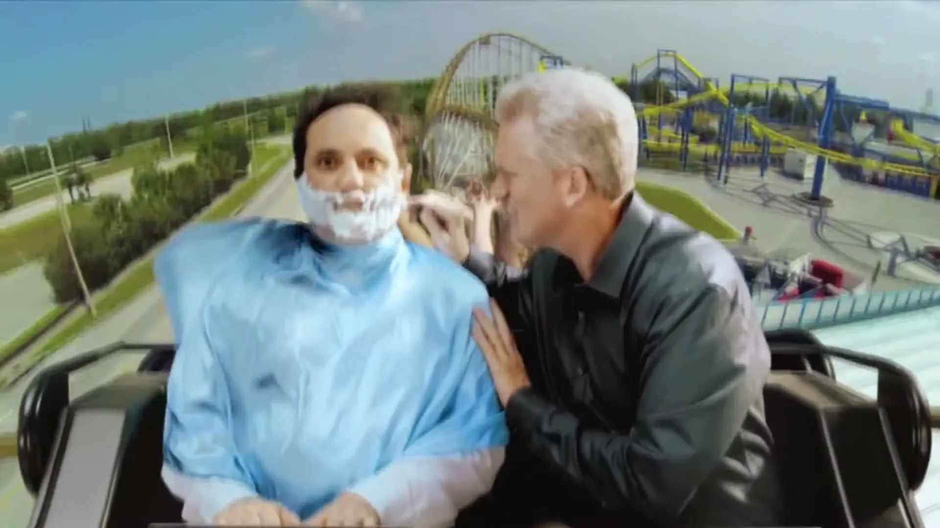 Two men on a rollercoaster, one with shaving cream on his face and a barber's cape, as the other shaves him with a straight razor.