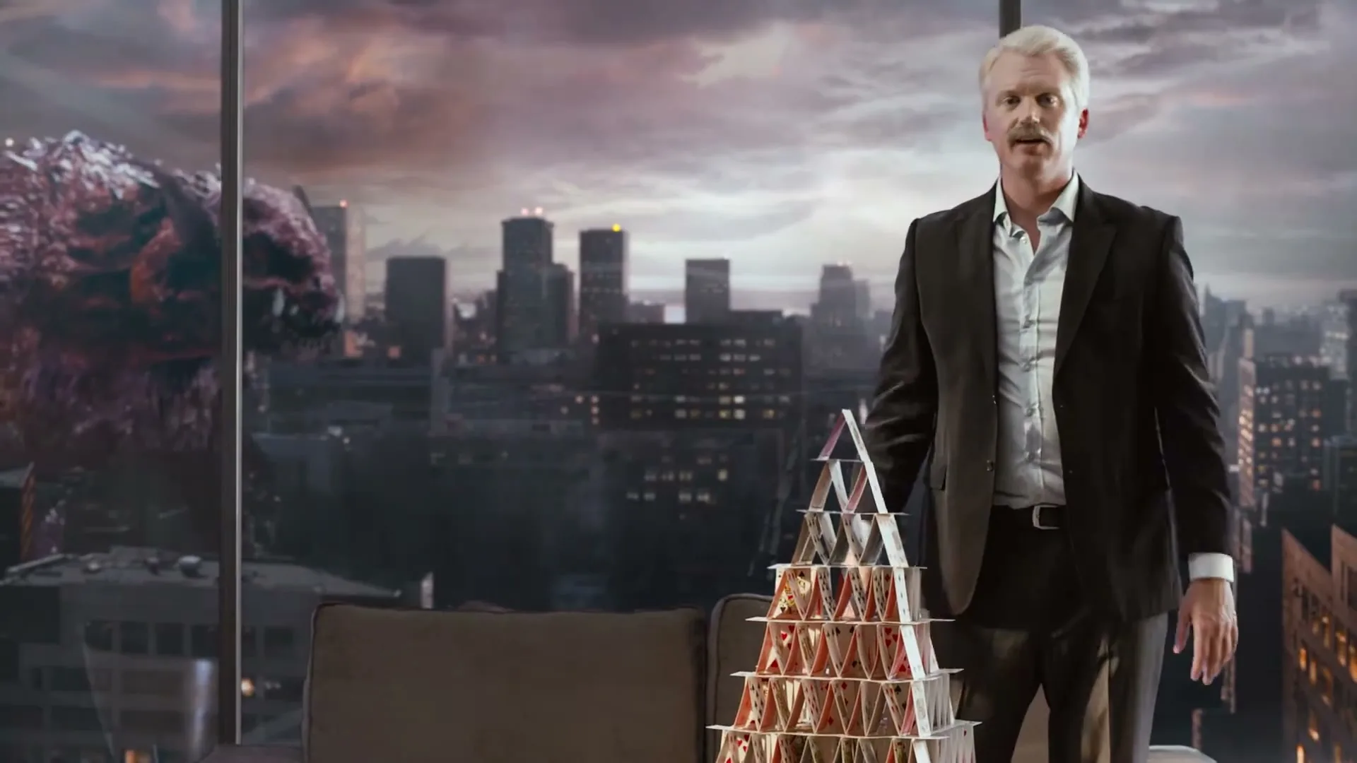 A man in a suit builds a house of cards in a high-rise apartment, with a giant monster visible outside the window in a city skyline.