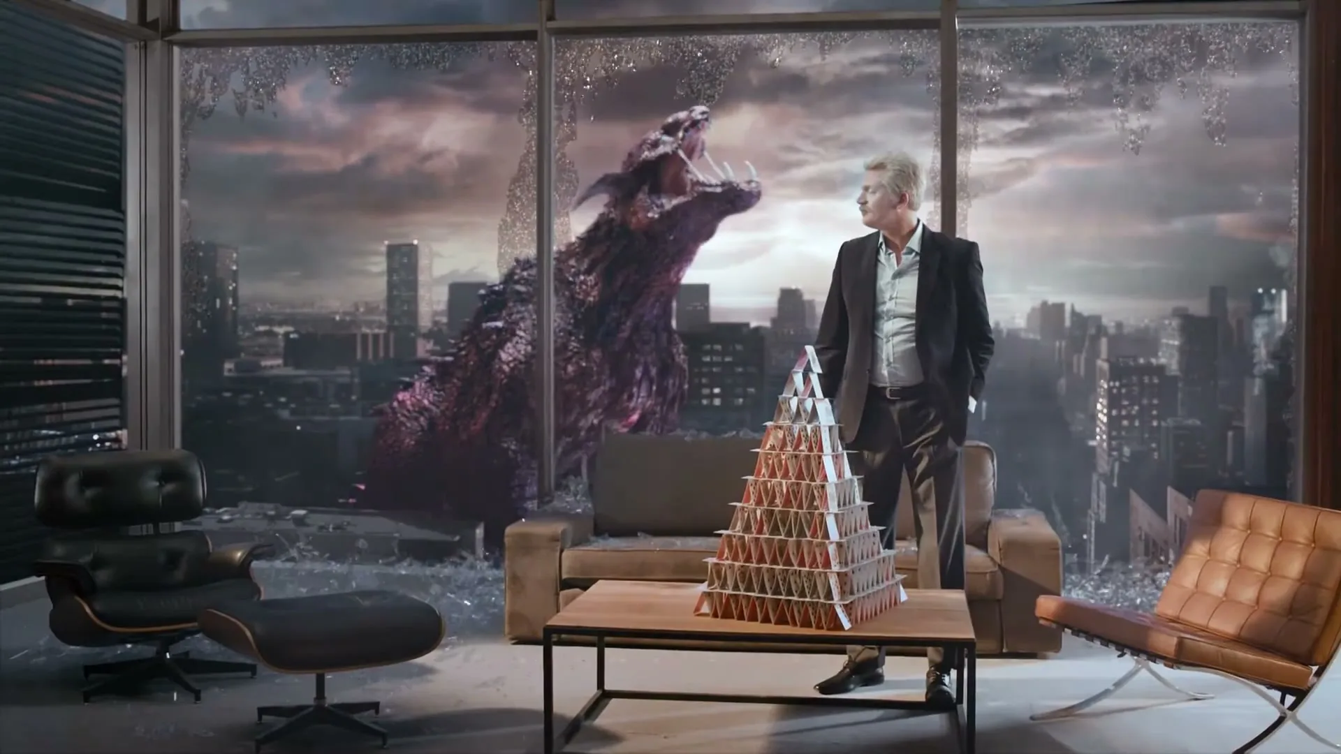 A man in a suit stands next to a house of cards on a coffee table, as a large purple monster roars outside a broken window overlooking a city.