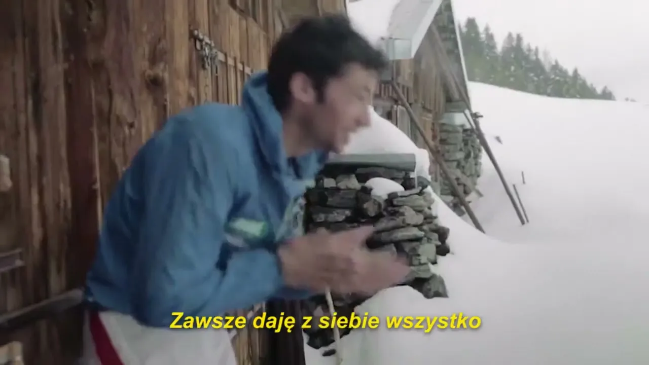 A male athlete in a blue jacket and white pants warms his hands outside a rustic wooden cabin in a snowy mountain landscape.