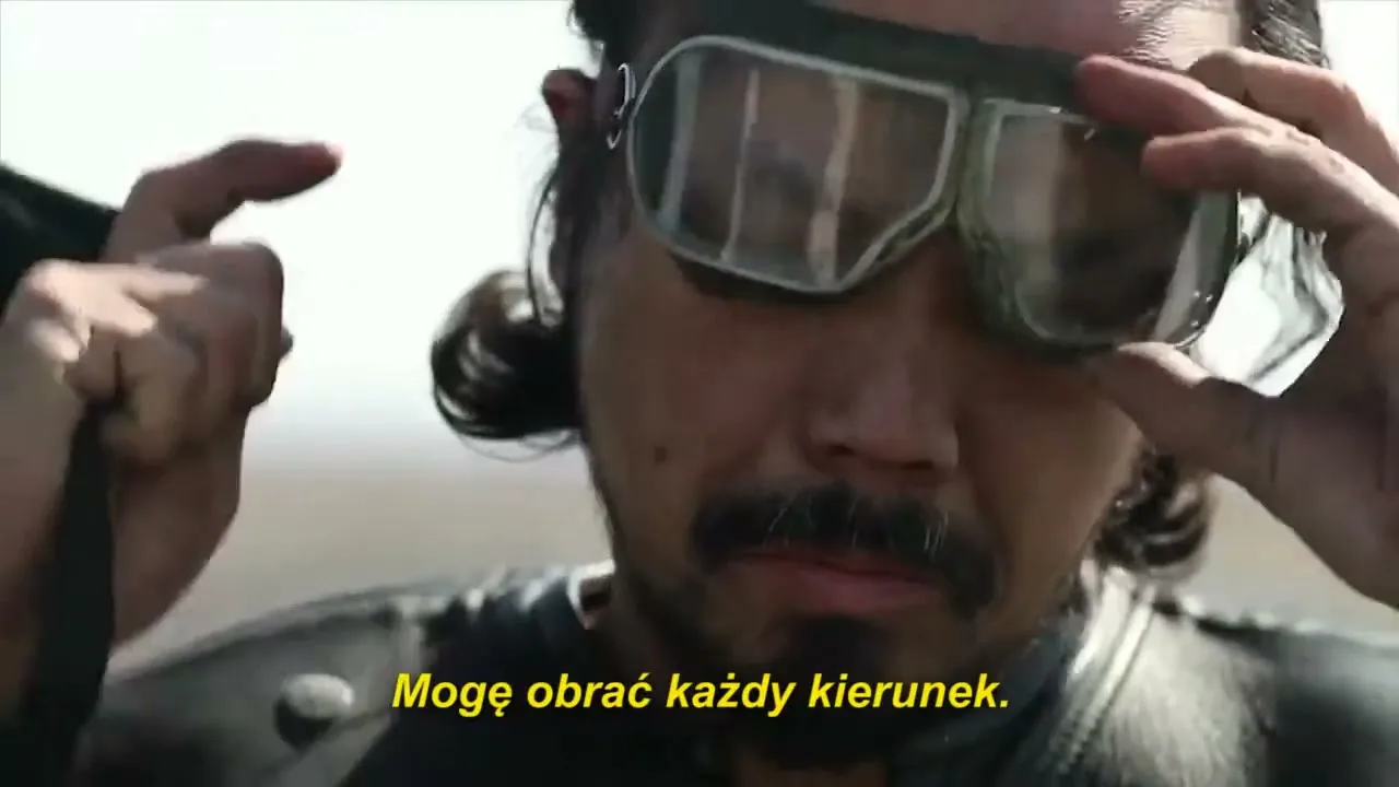 A man with long hair and a mustache adjusts his goggles on his head, looking down with a serious expression outdoors.