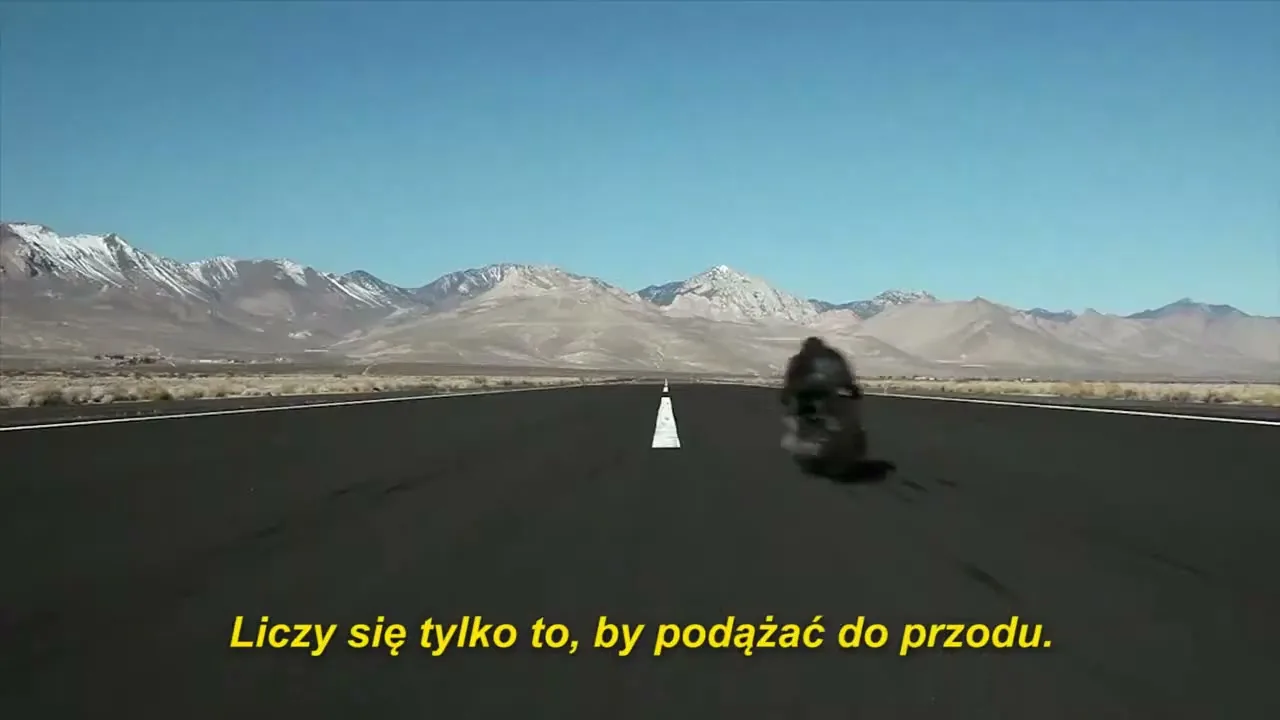 A lone motorcyclist speeds down an empty runway towards distant snow-capped mountains under a clear blue sky.
