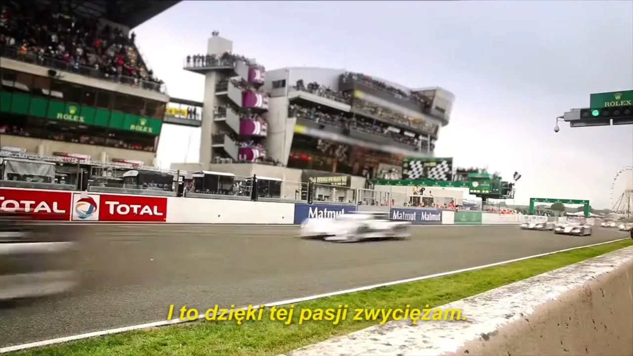 Racing cars speed past the camera on a track, with spectators in stands and buildings in the background under a bright sky.