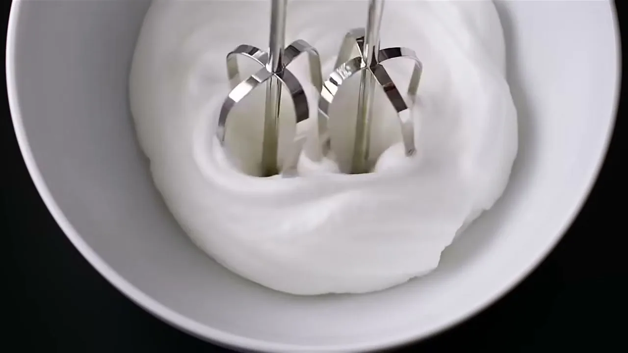 A hand mixer whips egg whites in a white bowl, transforming them from liquid to fluffy foam, in a kitchen setting.