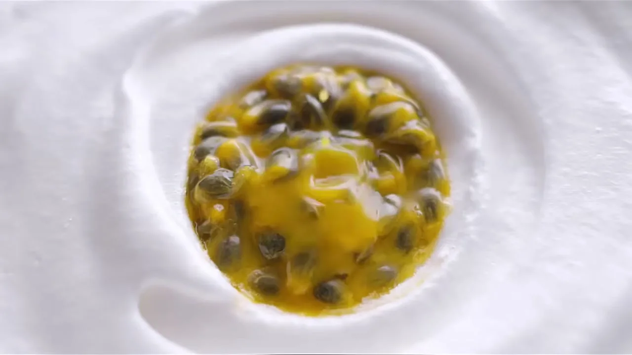 A close-up shot shows vibrant yellow passion fruit pulp with seeds, swirling in a white creamy substance, creating a visually appealing food shot.