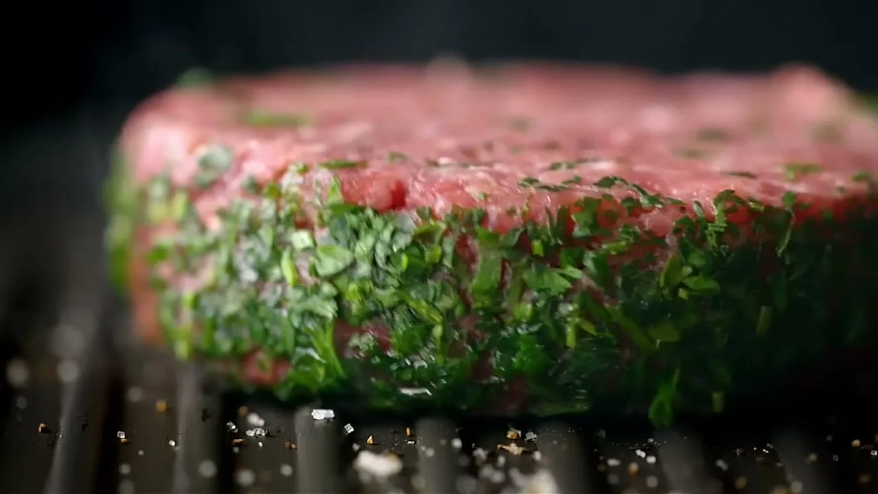 A raw, herb-crusted burger patty sizzles on a hot grill, with salt and pepper flakes scattered around it.