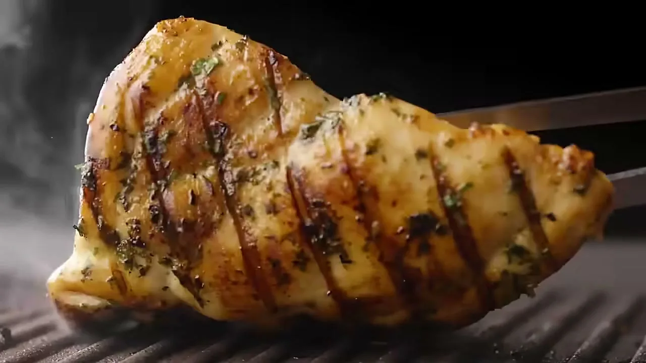 A close-up shot shows a piece of grilled chicken with herbs being turned over on a grill with tongs, emitting smoke.