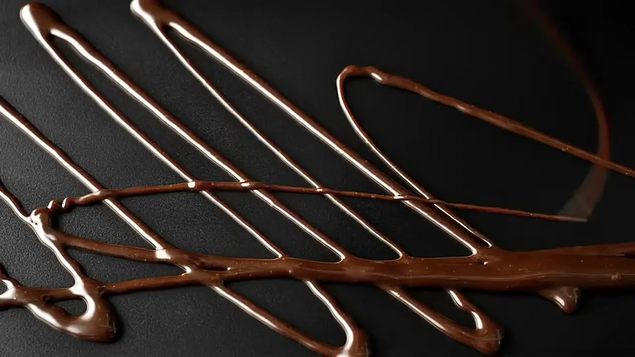 Melted chocolate is drizzled onto a dark surface, creating intricate patterns as it spreads and overlaps.