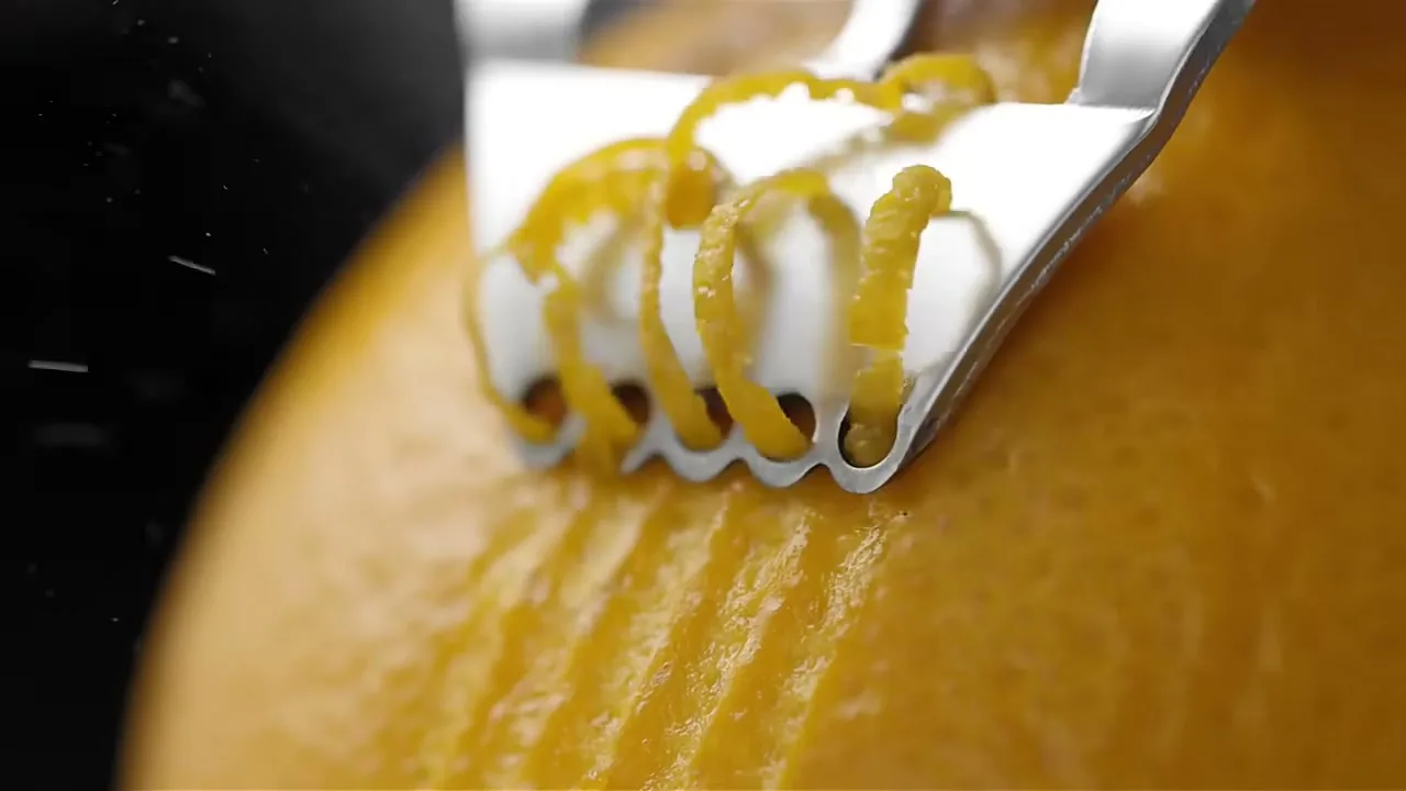 A close-up shot shows a metal zester peeling thin, curly strips of zest from a bright yellow lemon.