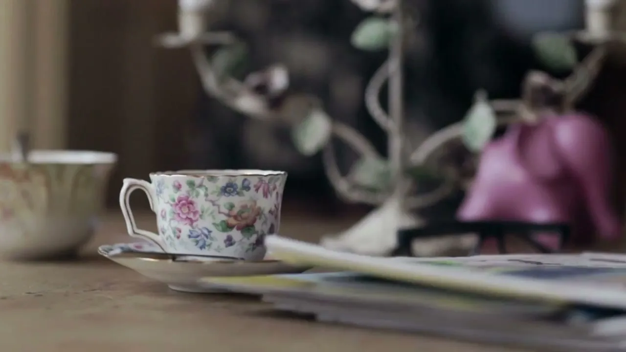 A floral teacup and saucer sit on a wooden surface, with a stack of newspapers landing next to it in a cozy interior setting.