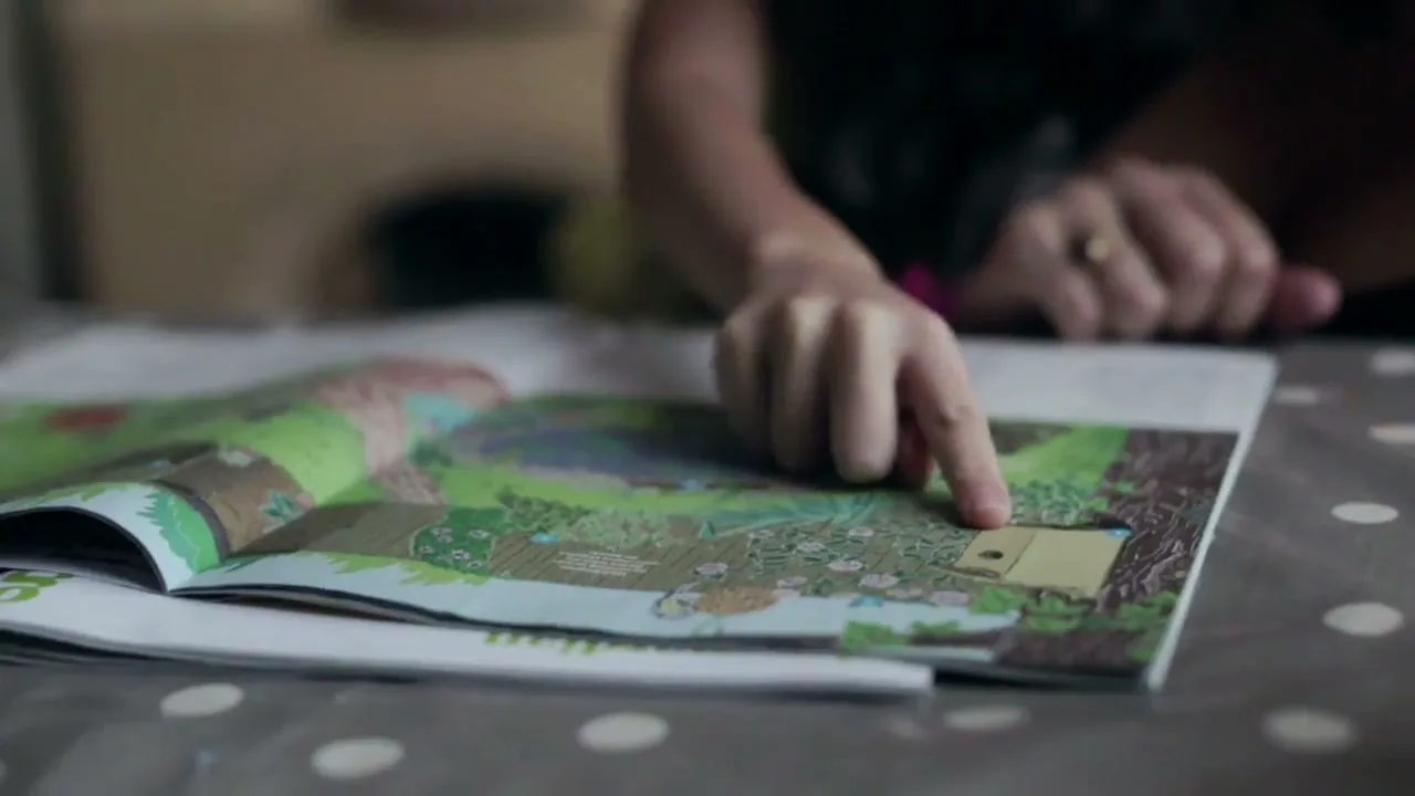 A person's hands point at a colorful children's book spread open on a polka-dotted table, showing a whimsical outdoor scene.