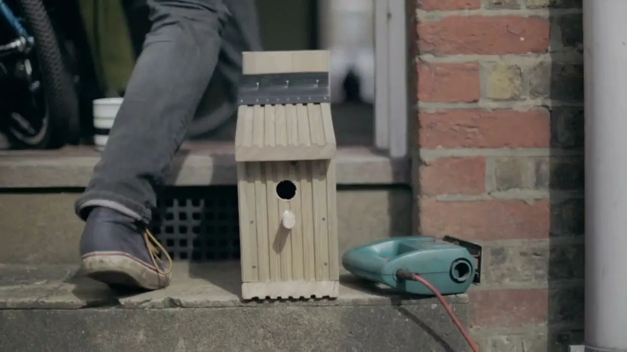 A person's legs and feet are visible next to a wooden birdhouse and a power tool on a concrete step outside a brick building.