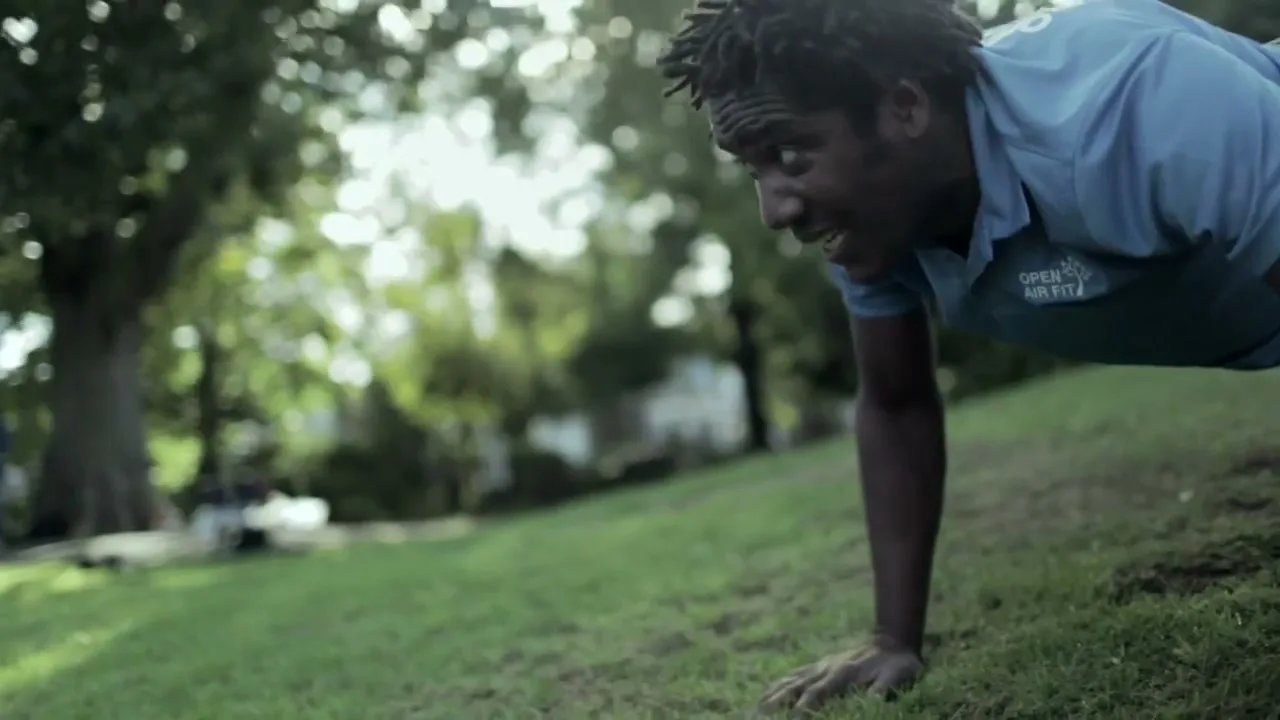 A man with dark skin and dreadlocks, wearing a blue polo shirt, does push-ups on a grassy hill in a park on a sunny day.