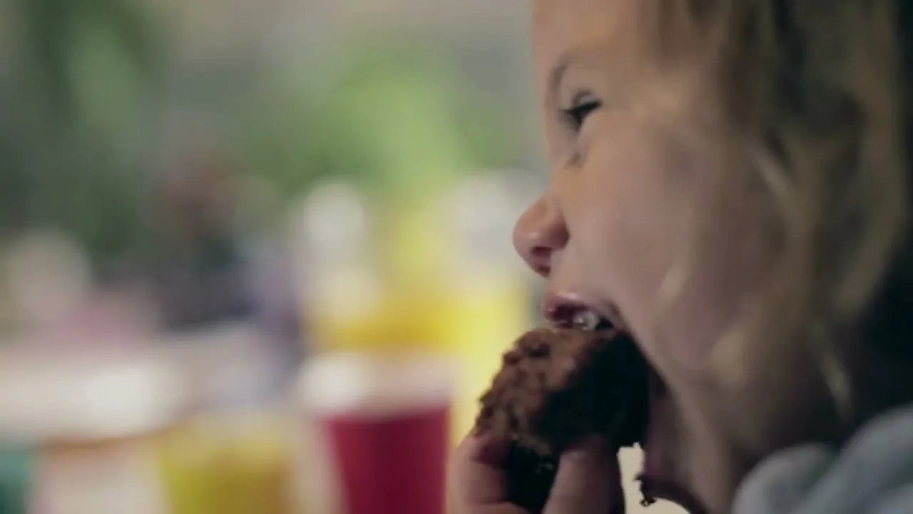 A young child with blonde hair eats a chocolate muffin, getting chocolate on their face, in a soft-focus interior setting.