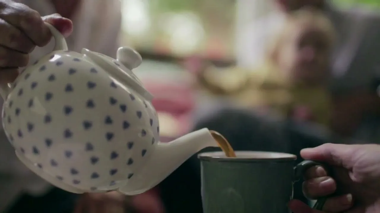 A person pours tea from a white teapot with blue hearts into a green mug, with blurred people in the background.