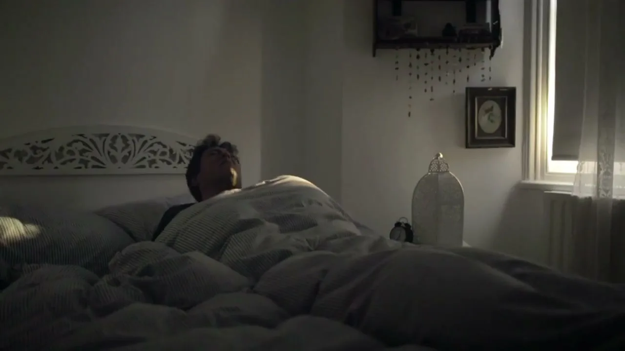 A man with dark, curly hair lies in bed, covered by a striped duvet, as sunlight streams through a window in a dimly lit bedroom.