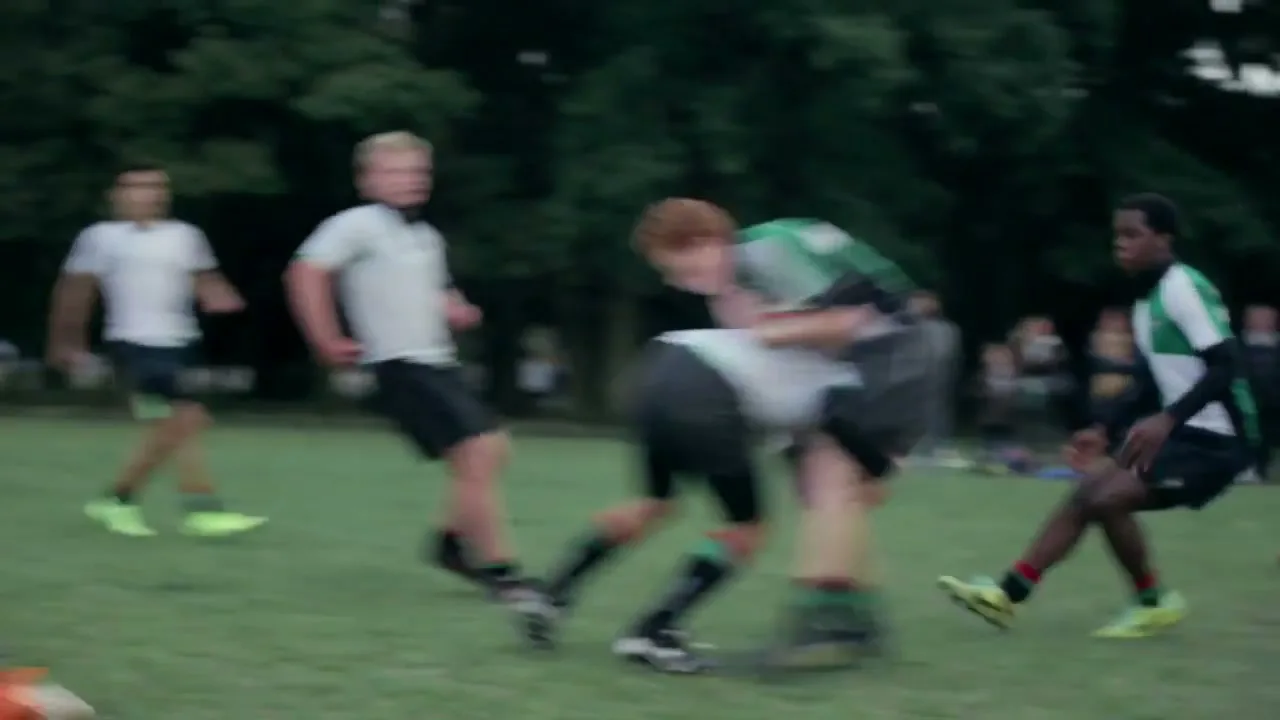 A red-haired teen boy in a green and white rugby shirt runs with a rugby ball on a grassy field, while other players chase him.