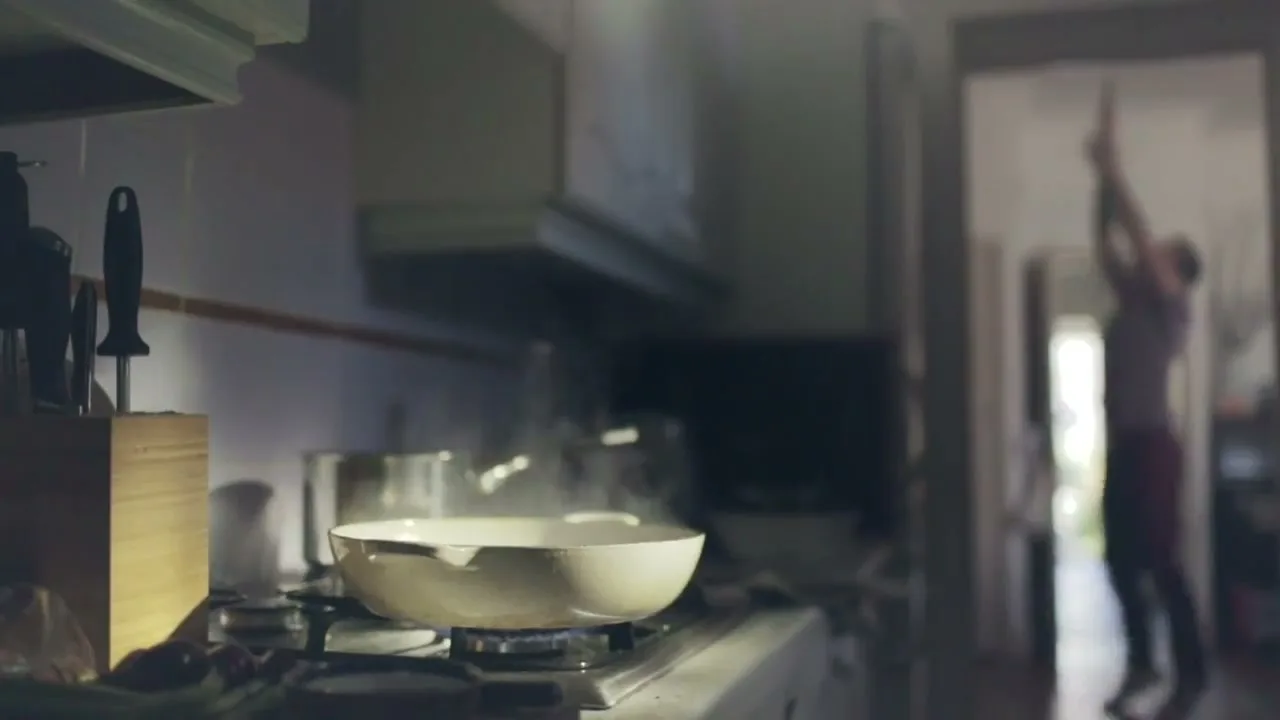 A steaming pan sits on a gas stove in a kitchen, while a person stretches in a doorway in the background.