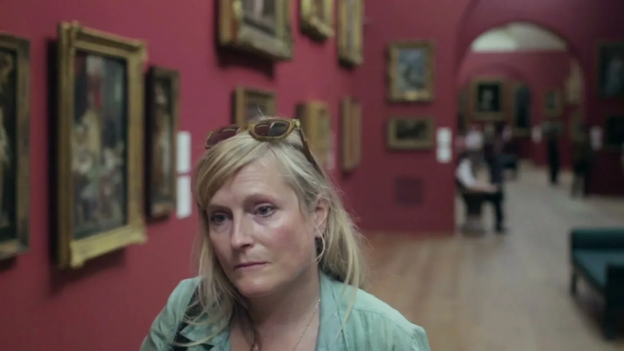 A blonde woman with sunglasses on her head looks around a museum gallery with red walls and paintings, appearing thoughtful.