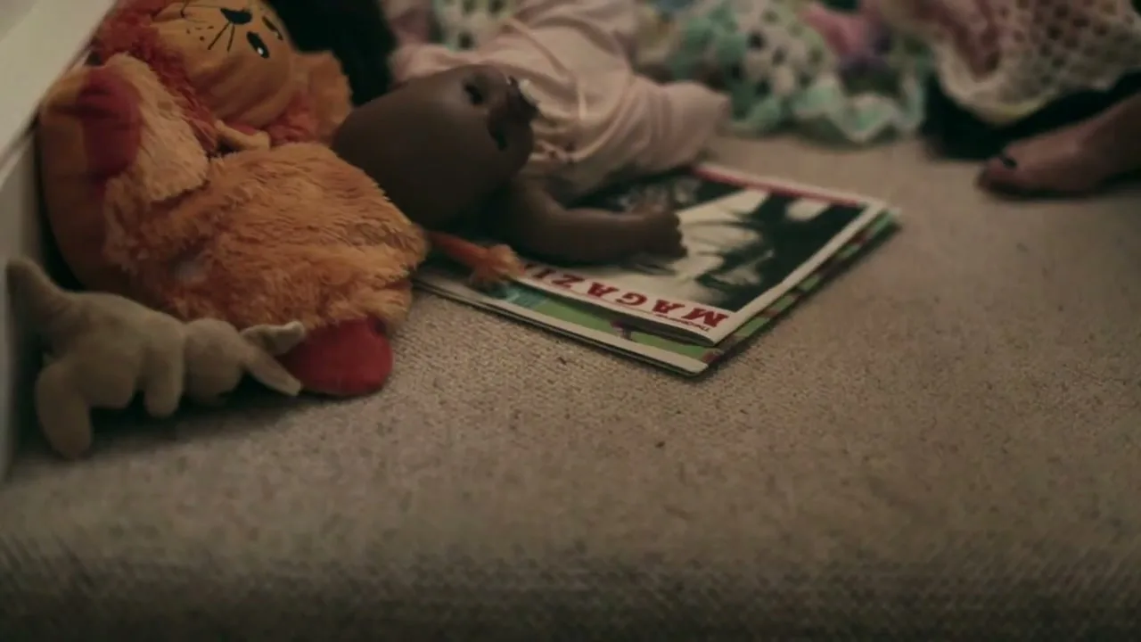 A woman's hand reaches down to pick up a magazine from a carpeted floor, surrounded by children's toys and blankets.