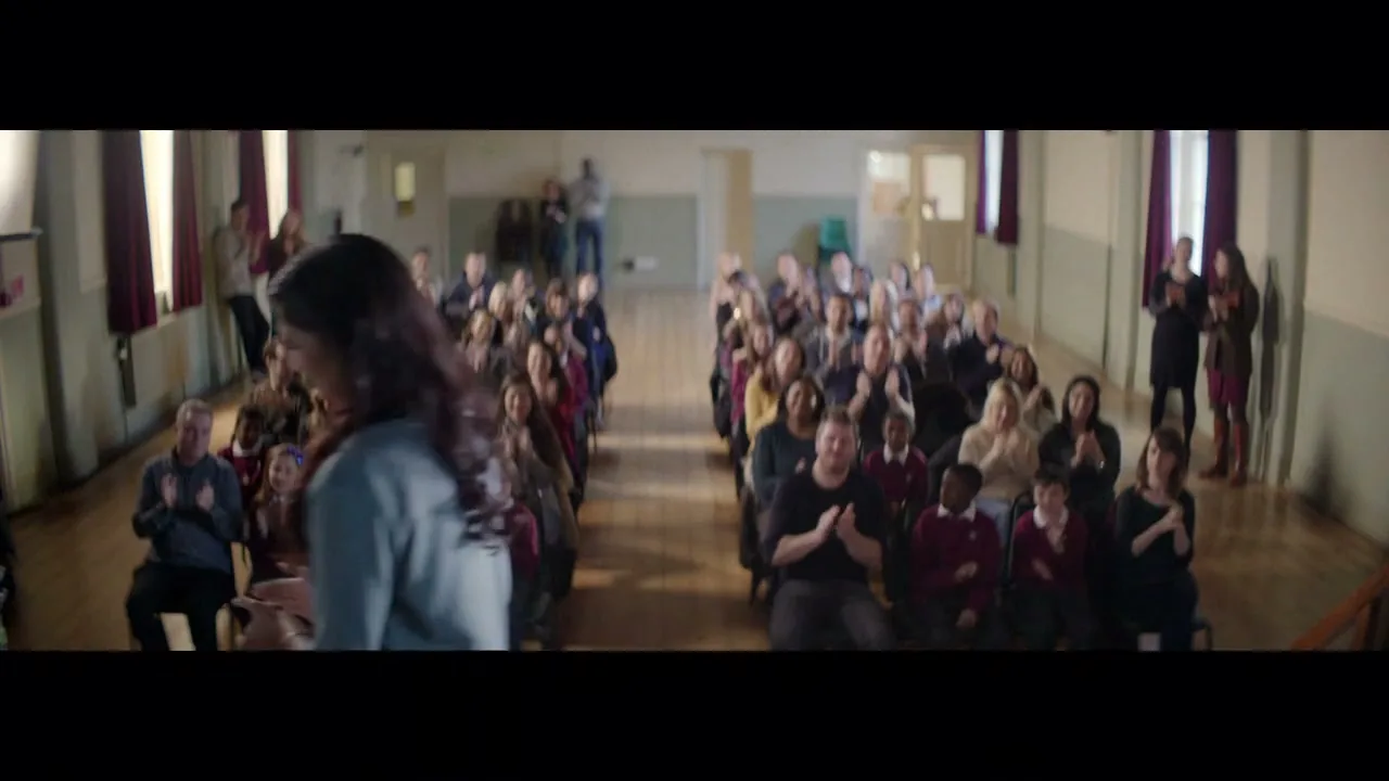 A young woman with long dark hair, wearing a grey cardigan, stands in front of a large audience of adults and children who are all clapping in a school hall.