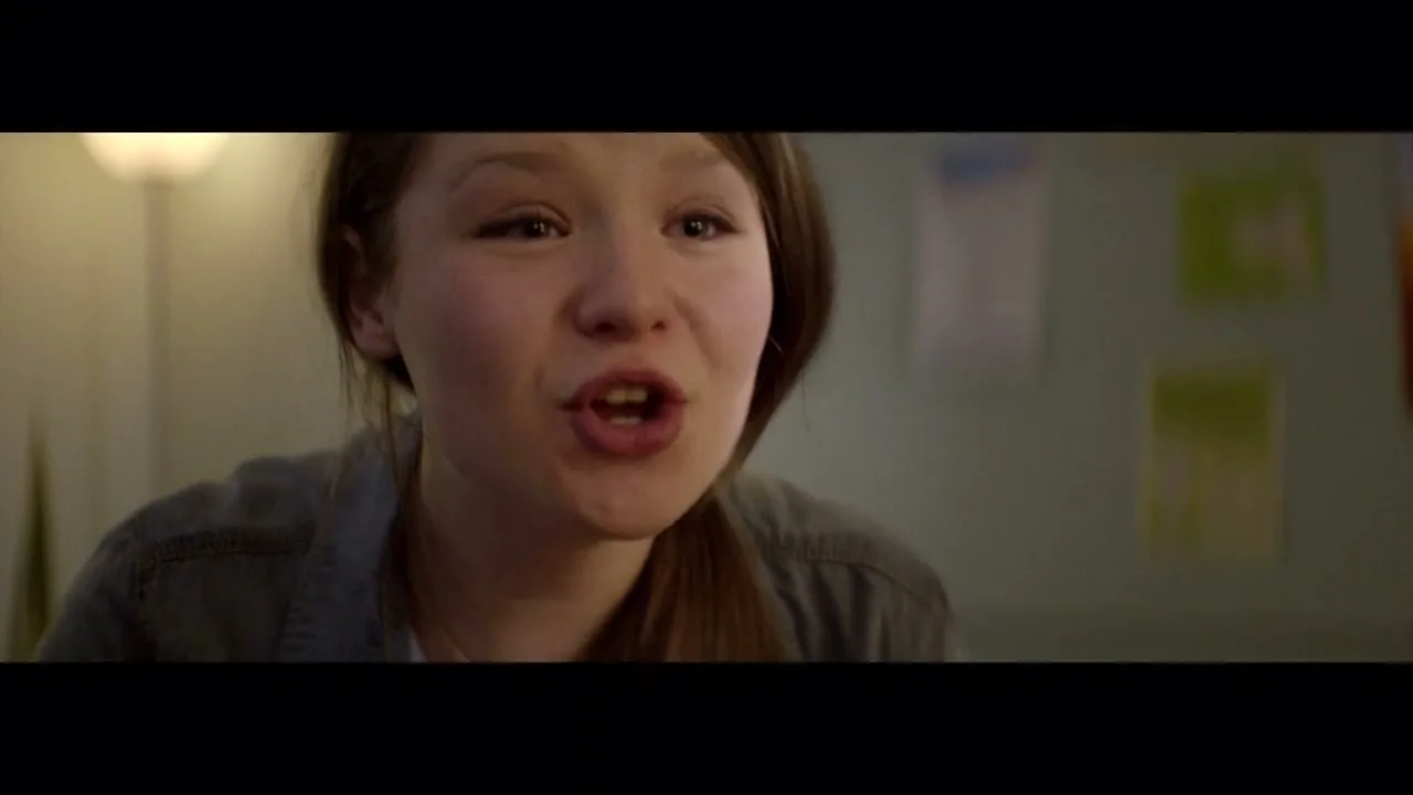 A young girl with long brown hair and a denim shirt looks distressed and speaks directly to the camera in a dimly lit room.