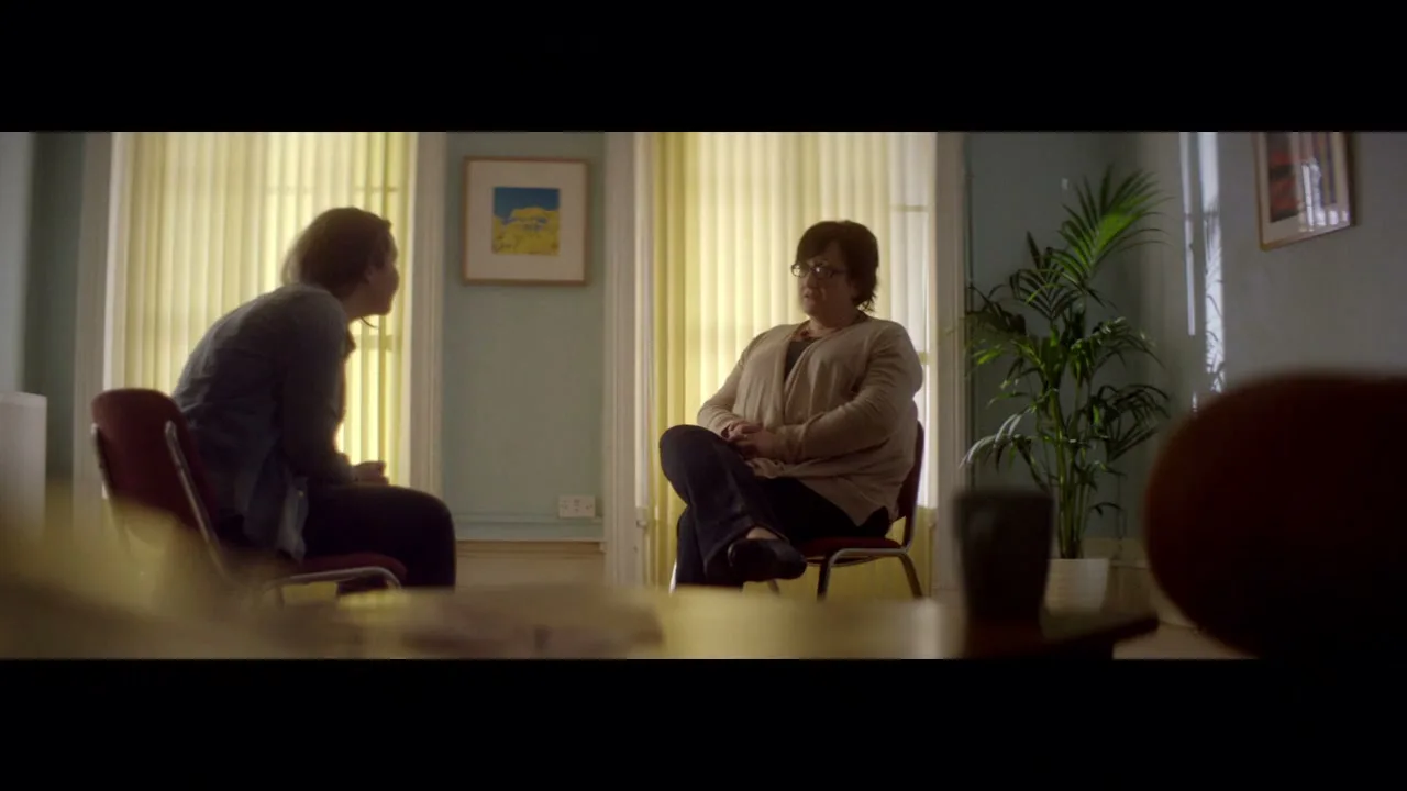 A young person with long hair sits opposite an older woman in a bright room, engaged in a serious conversation.