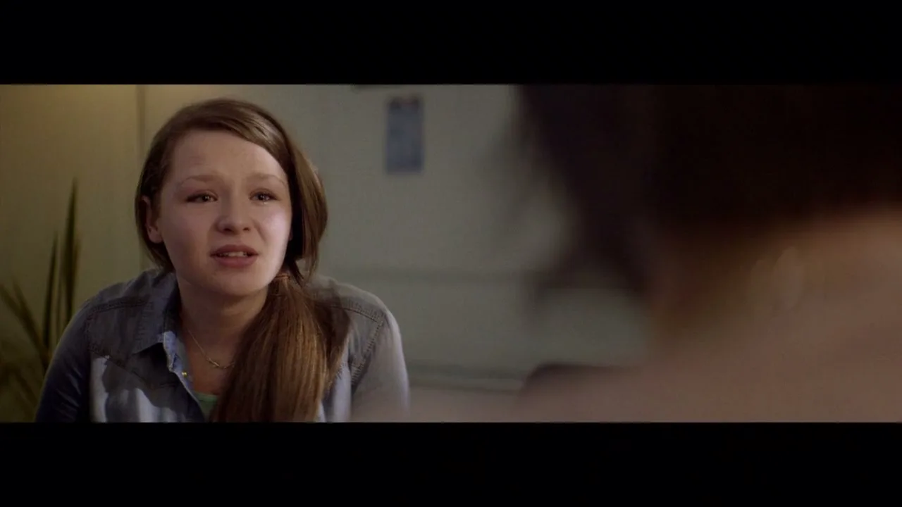 A young girl with long brown hair and a denim shirt looks distressed while talking to someone off-screen in an interior setting.