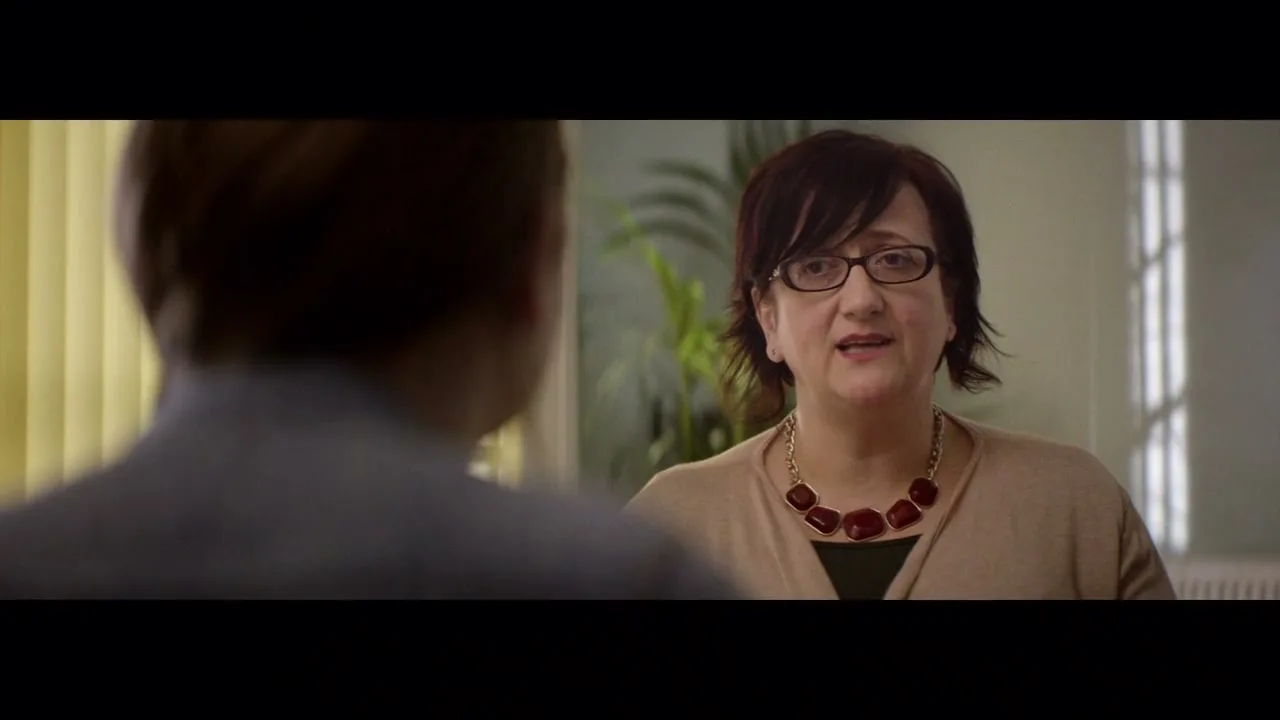 A female social worker with dark hair and glasses, wearing a beige cardigan and a red necklace, speaks to a person whose back is to the camera in an office.