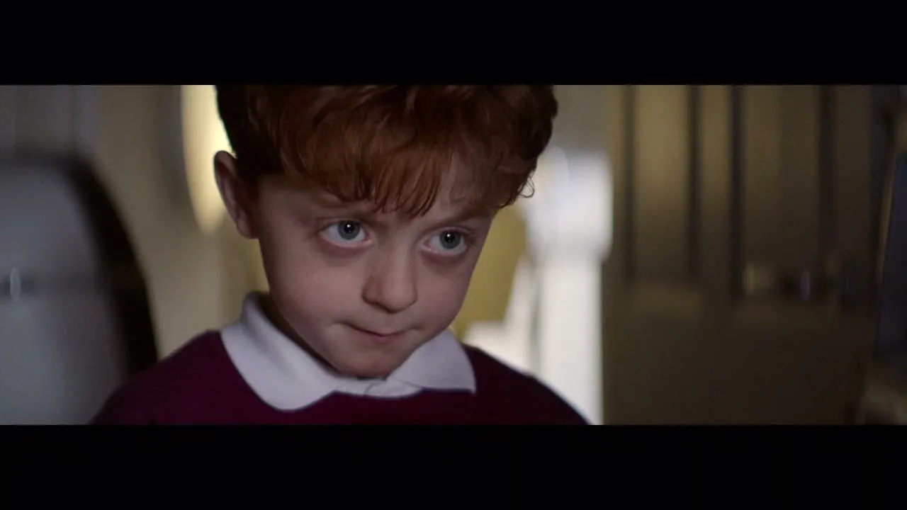 A young boy with red hair and a white collar looks down with a worried expression in a dimly lit interior.