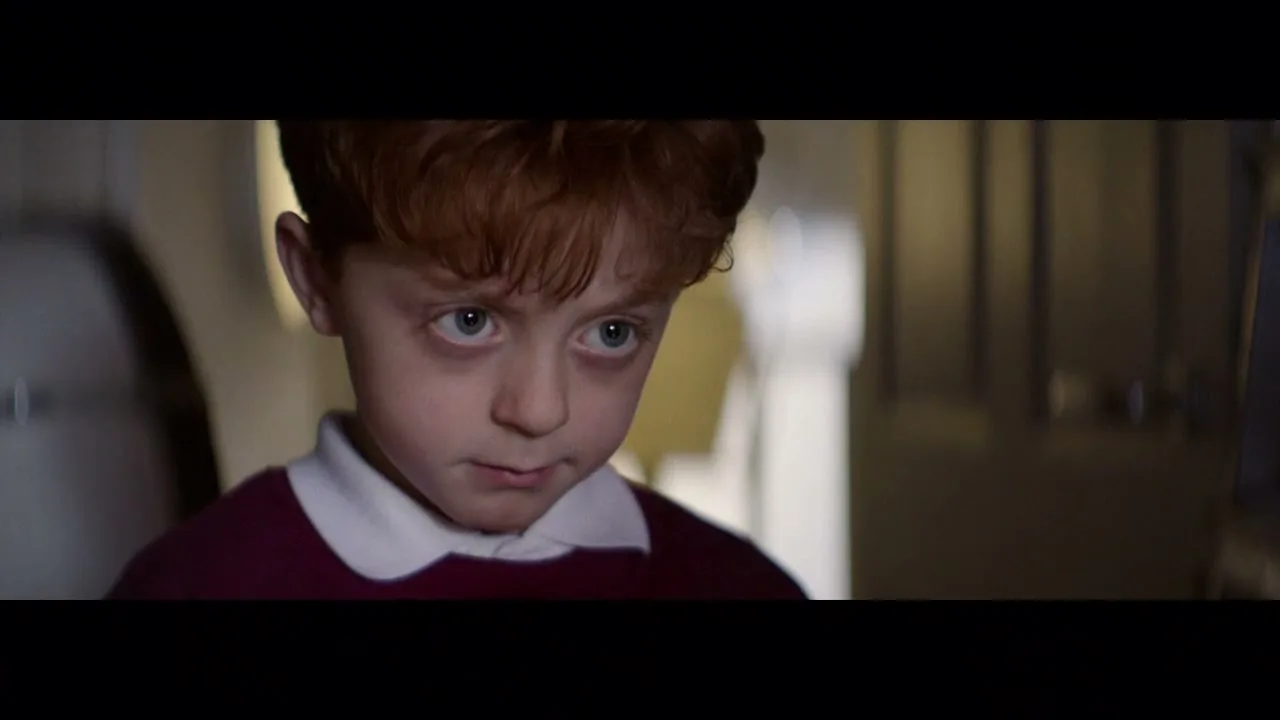 A young boy with red hair and a white collar shirt looks directly at the camera with a worried expression in a dimly lit room.