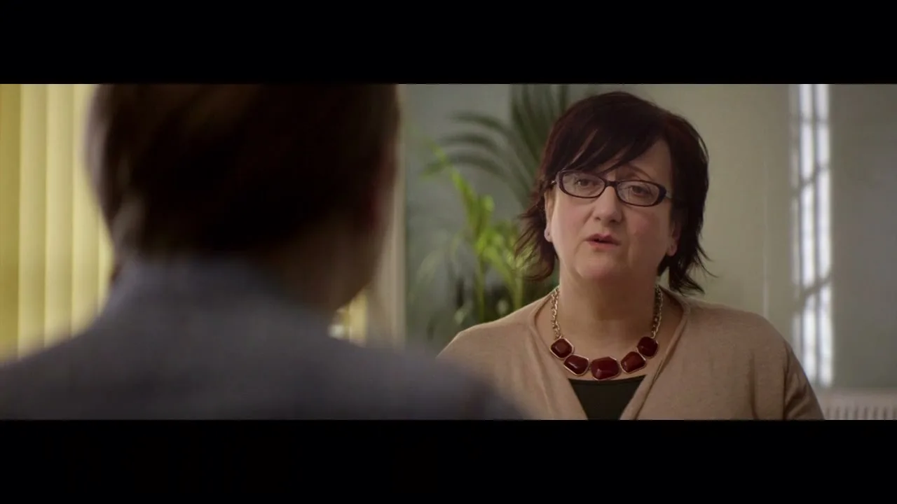 A female social worker with dark hair and glasses talks to a young person, seen from behind, in an office setting.
