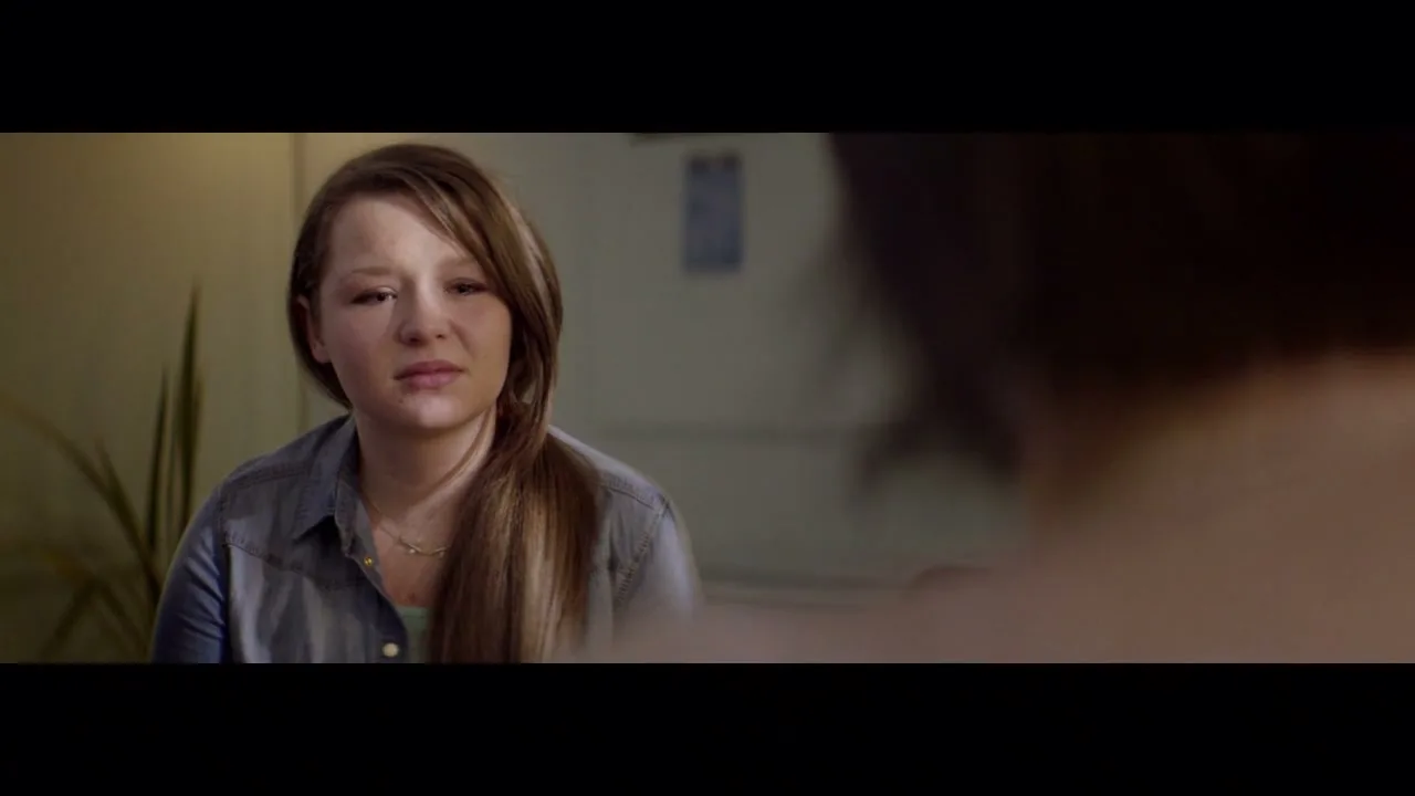 A young woman with long brown hair, wearing a denim shirt, looks directly at the camera with a sad expression in an interior setting.