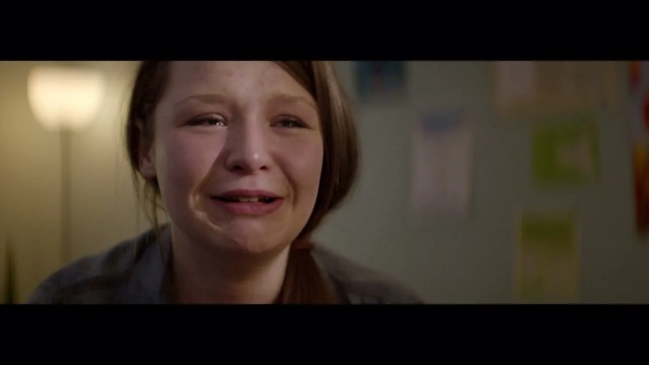 A young girl with brown hair cries and speaks, her face illuminated by a lamp in a dimly lit room with drawings on the wall.