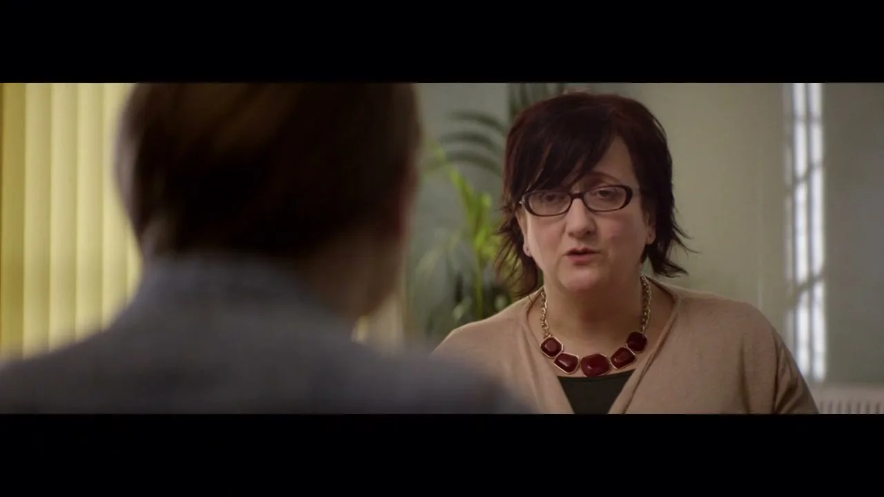 A female social worker with dark hair and glasses talks to a person whose back is to the camera, in an office setting.