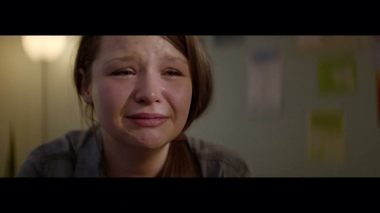 A young woman with long brown hair, wearing a grey shirt, cries with her eyes closed in a dimly lit room.
