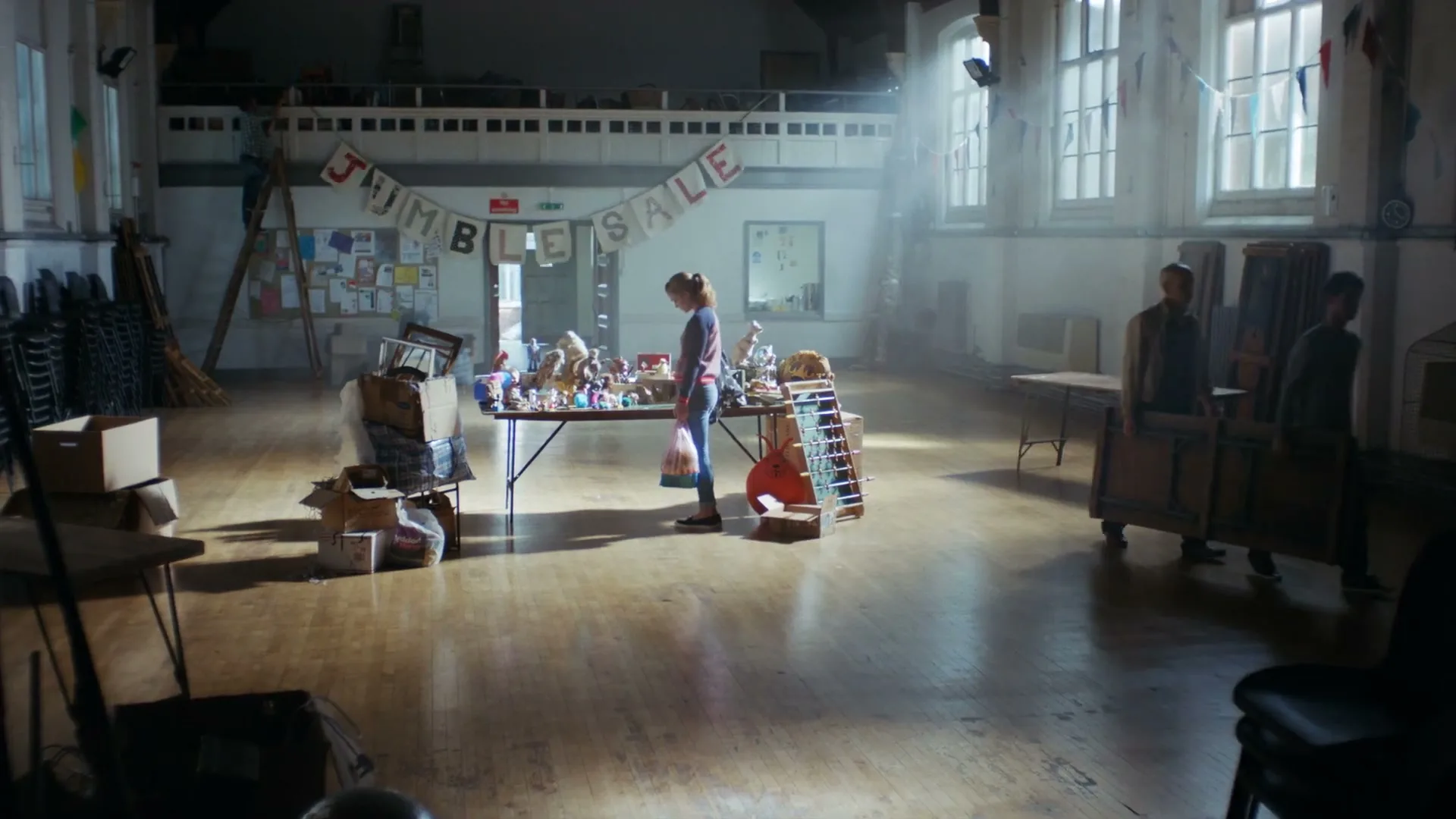 A young woman stands alone at a table filled with items, while two men carry a large wooden box in a dusty, sunlit community hall.