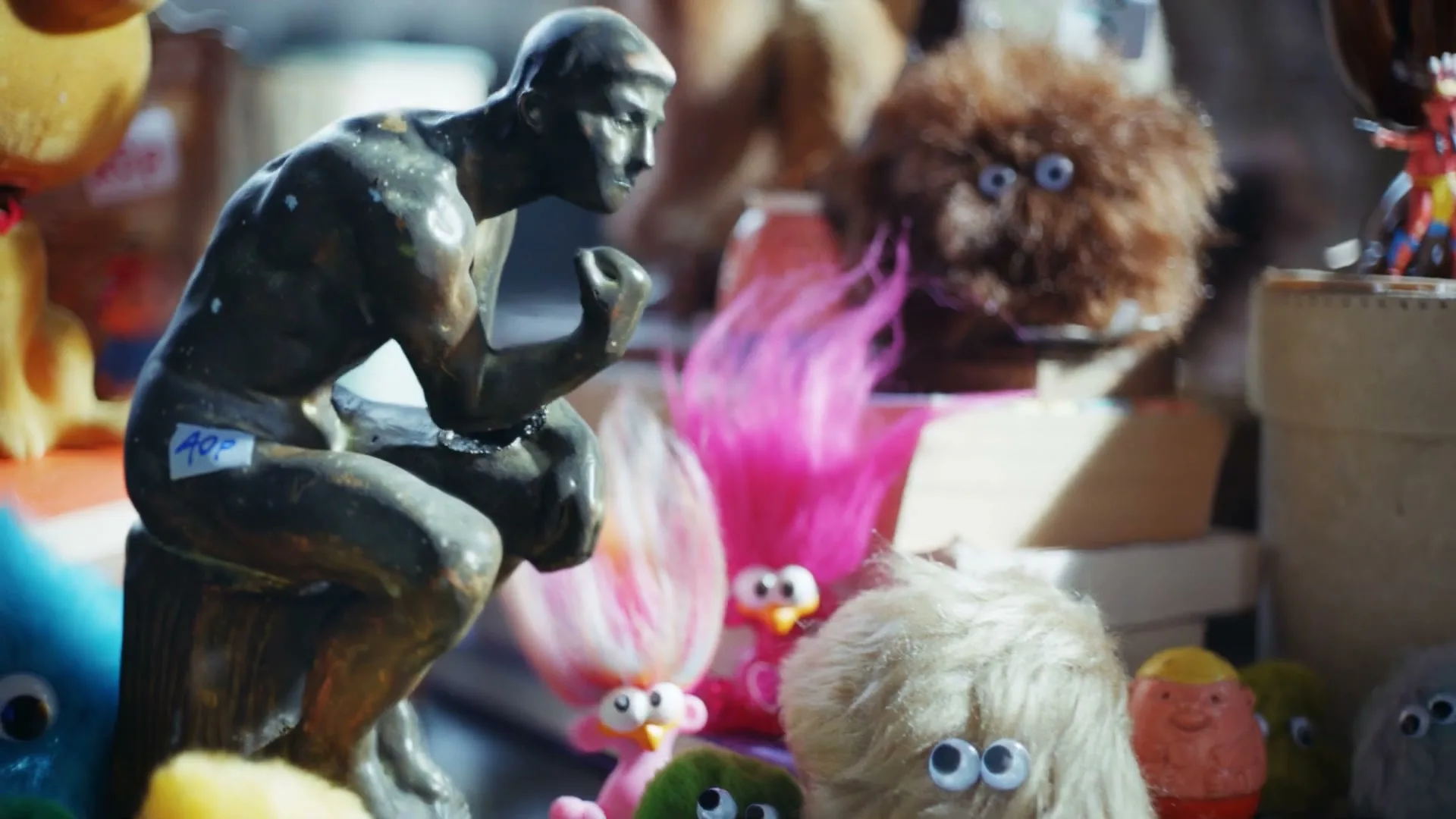 A person places a worn teddy bear on a table full of old toys and trinkets, with a cardboard sign reading "UNSOLD (PLEASE TAKE)" in a dimly lit room.