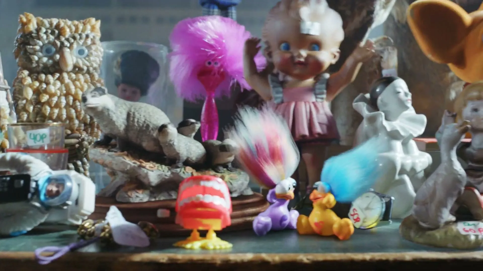 A wide shot of a table full of various discarded toys and trinkets, including a teddy bear, dolls, and action figures, in a dusty room.