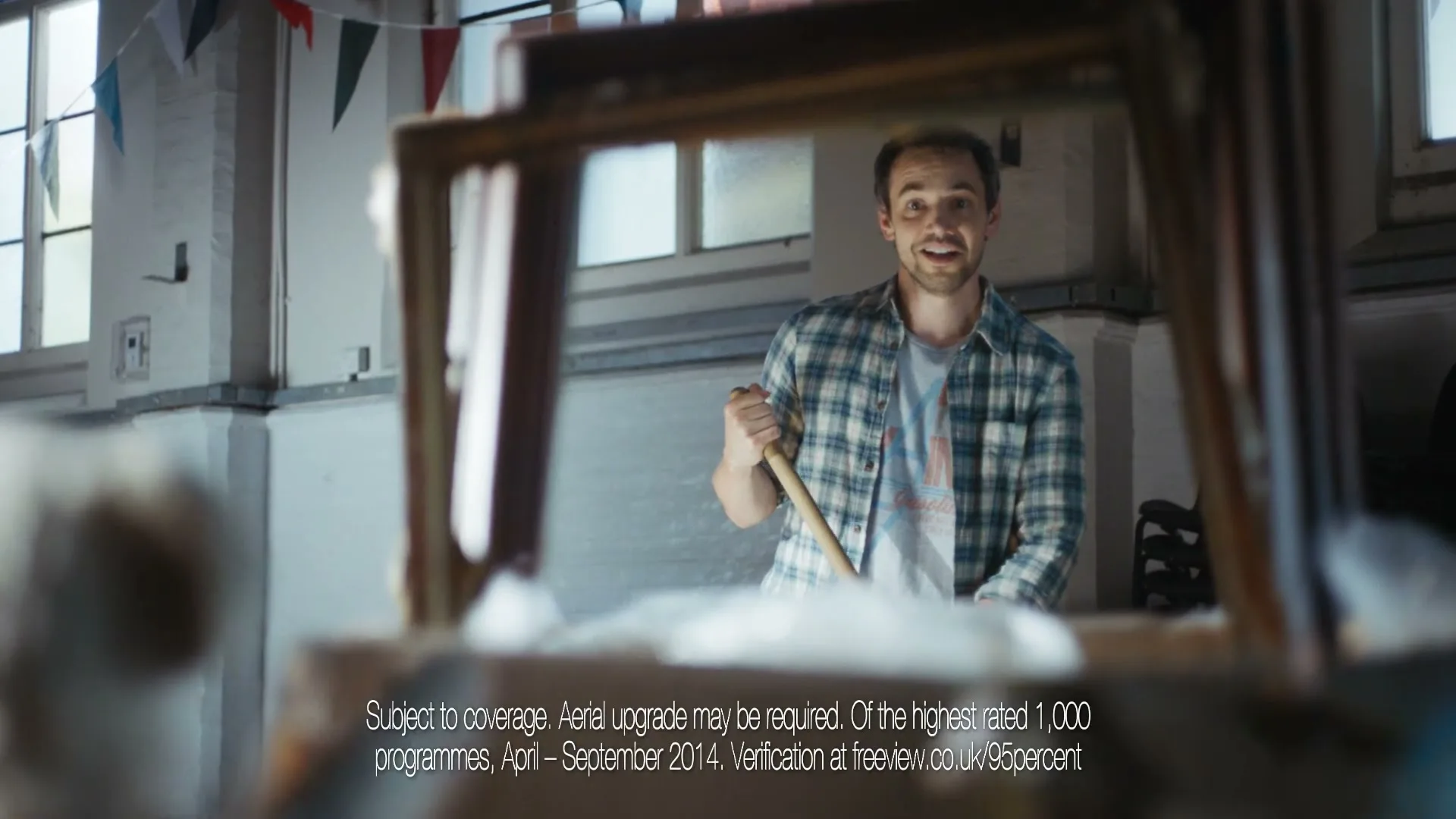 A young man in a plaid shirt and t-shirt smiles while holding a broom, seen through a blurry foreground in a large, bright room.