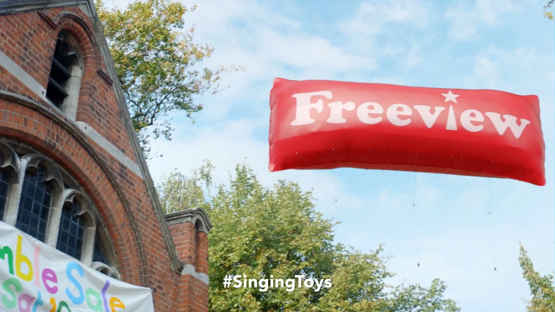 A red inflatable balloon with "Freeview" written on it floats in the sky above a brick building with a "Jumble Sale" banner and trees.
