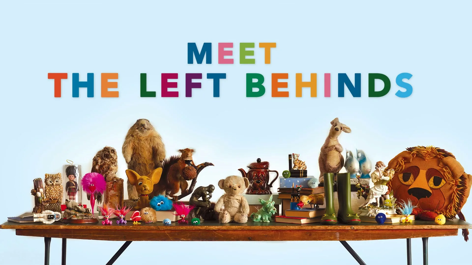 A wide shot of a collection of forgotten toys and objects on a wooden table, with the text "MEET THE LEFT BEHINDS" and "#SingingToys" displayed.
