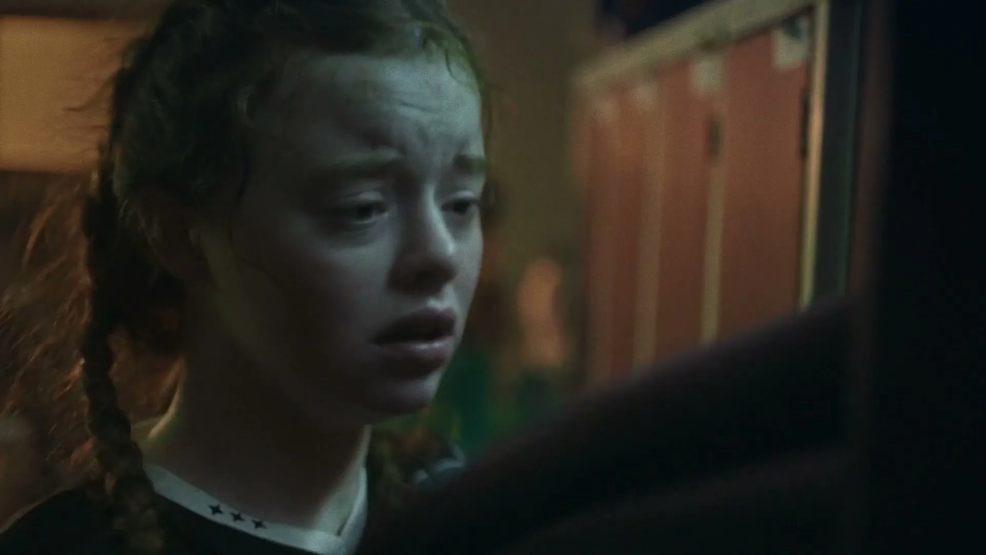 A young female soccer player with braided hair, wearing a green and black jersey, looks sad and wipes her face in a locker room.