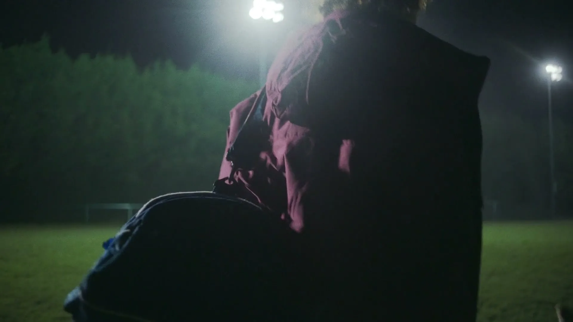 A young female athlete in a maroon jacket walks across a misty green sports field at night, carrying a hockey stick and bag.