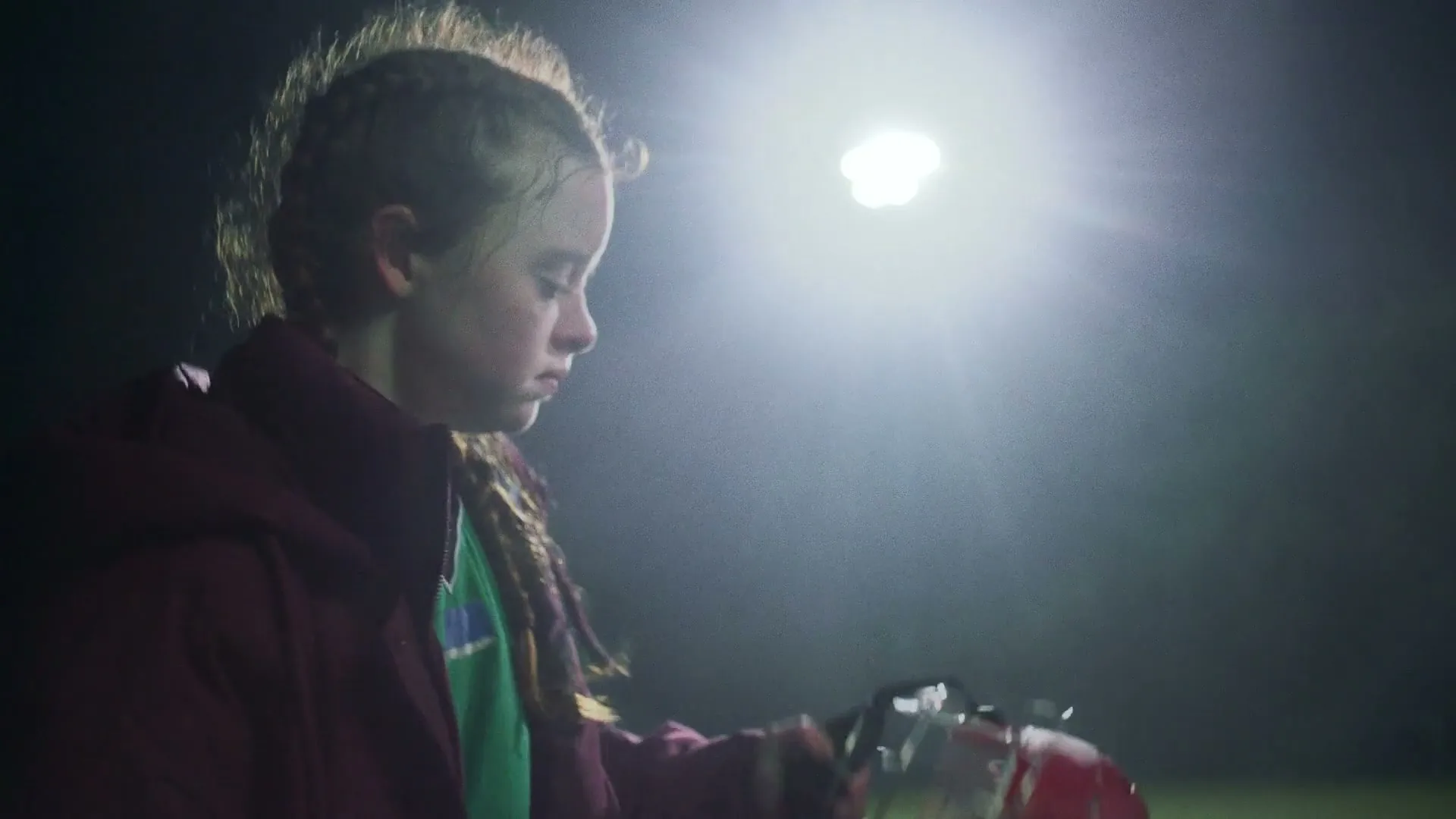 A young female athlete in a green jersey and maroon jacket looks down, holding a red helmet under bright stadium lights at night.