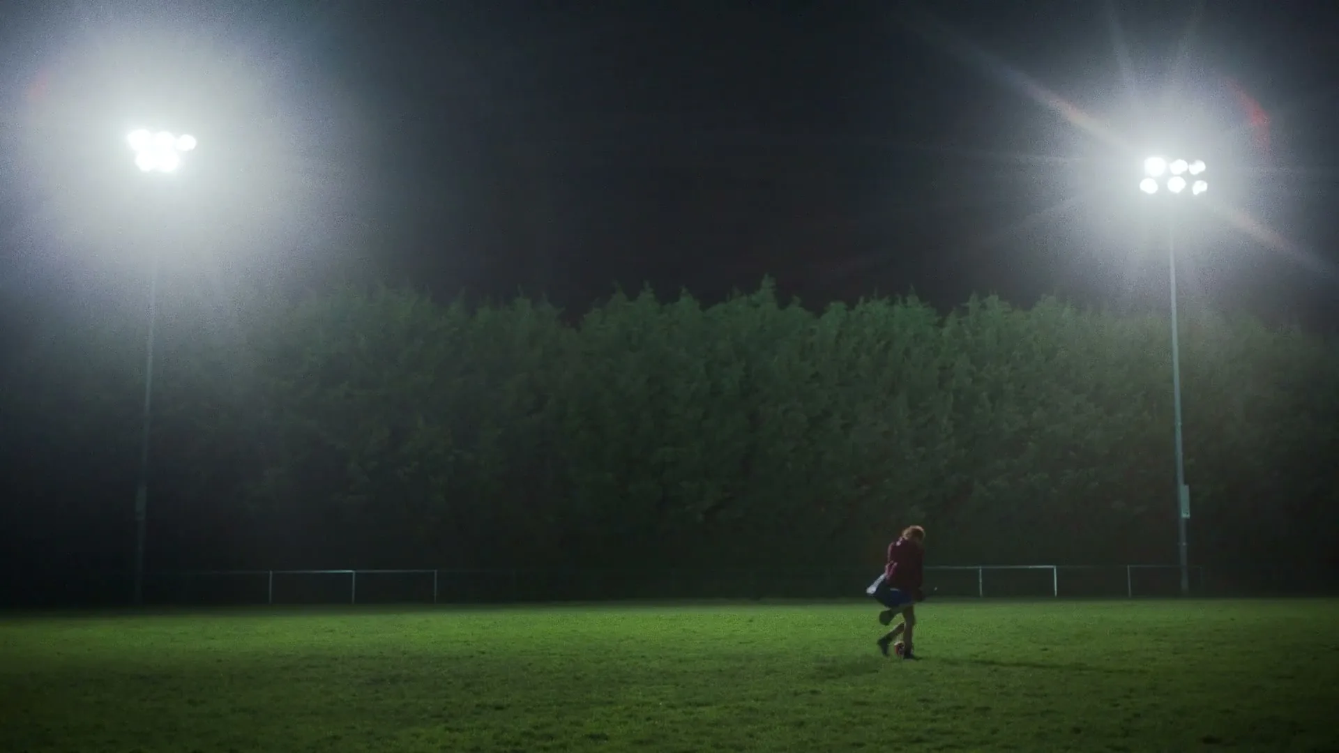 A young female athlete in a red jacket and blue skirt walks across a floodlit sports field at night, carrying a sports bag.