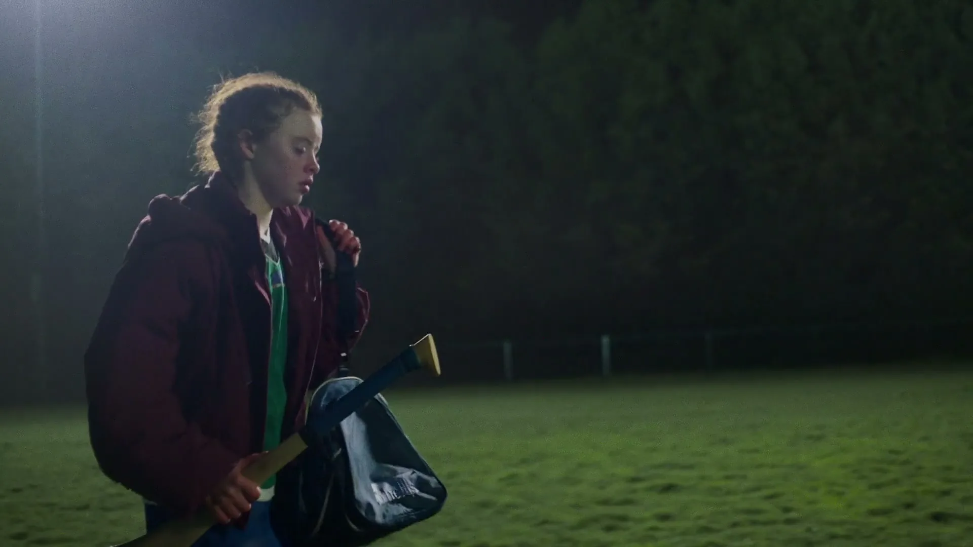 A young female athlete, dressed in a maroon jacket and blue shorts, walks across a dark sports field at night, carrying a hurley stick and a bag.