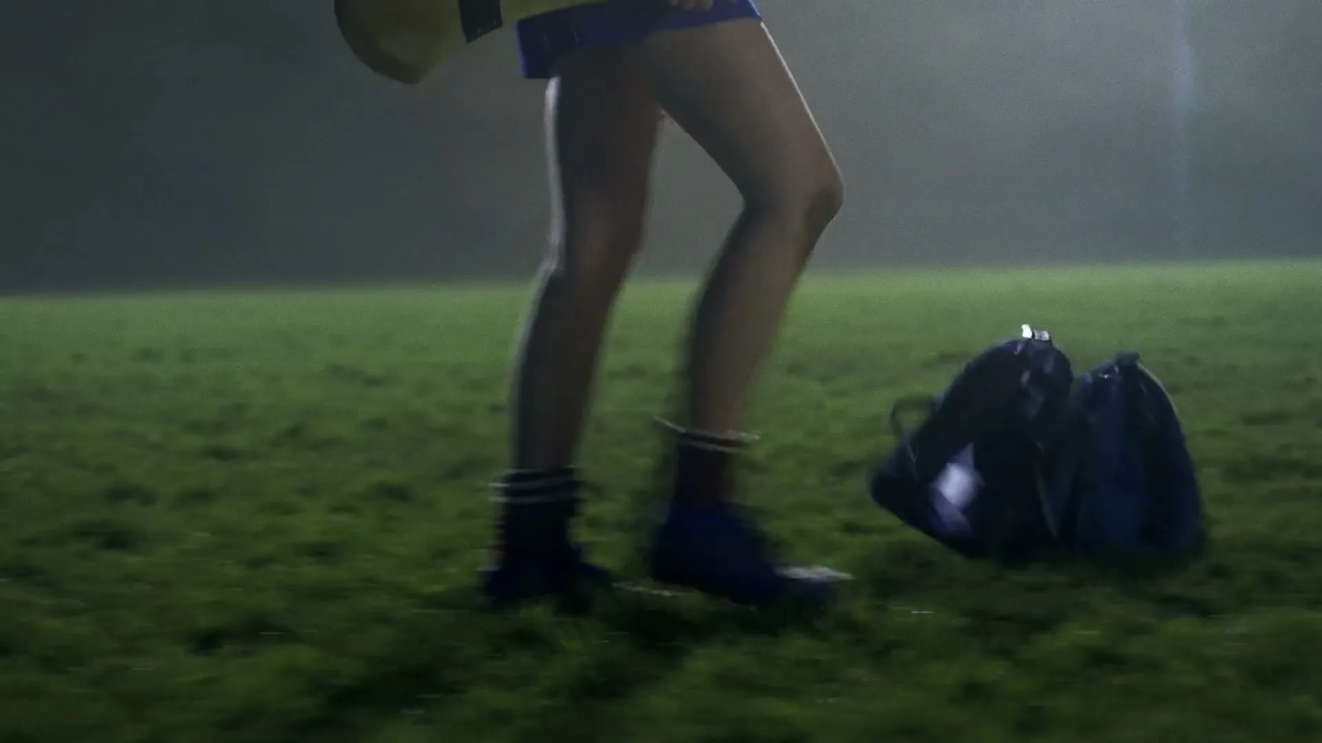 A young female athlete in a blue uniform and cleats walks across a grassy field at night, dropping a blue drawstring bag.
